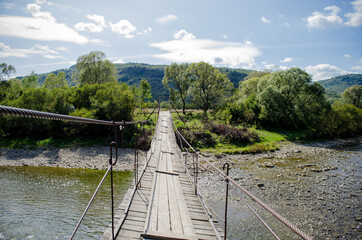 Fototapeta premium View of a rocky shallow mountain river from a tall old wooden suspension bridge in sunlight