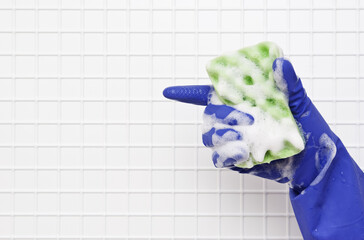 Hand in blue glove finger green small sponge in the foam on a background of white tiles