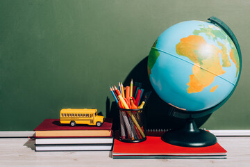 Globe and pen holder on notebook near school bus model on books near green chalkboard