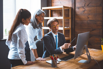 Team work of diverse office team. Young brunette and gray haired asian woman listen their male colleague pointing to computer monitor. Coworking concept. Tinted image.