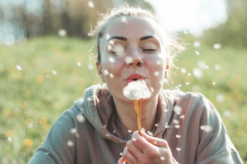 Fototapeta premium Young spring fashion woman blowing dandelion in spring garden. Springtime. Trendy girl at sunset in spring landscape background. Allergic to pollen of flowers. Spring allergy