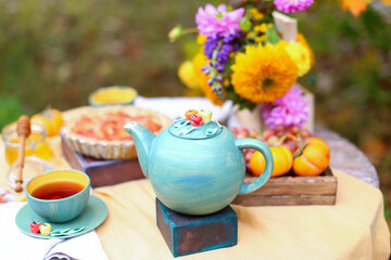 Autumn picnic. Tea party with beautiful kettle, cups at wooden table in garden. Harvest festival. Honey with stick, spoon, apple pie, persimmons, grapes, maple leaf, flowers, yellow linen tablecloth