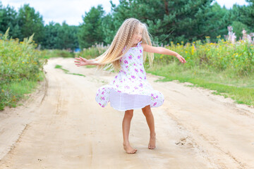 A girl with long blond curly hair is dancing, spinning on a forest road. Childhood, happiness, freedom of movement, dancing, reuniting with nature. Beautiful summer landscape.