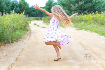 A girl with long blond curly hair is dancing, spinning on a forest road. Childhood, happiness, freedom of movement, dancing, reuniting with nature. Beautiful summer landscape.