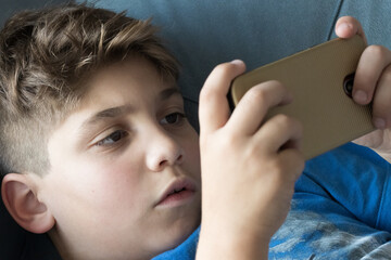 Menino deitado no sofá segurando o celular com as mãos e jogando. Fazendo aula online. Se divertindo.  - obrazy, fototapety, plakaty