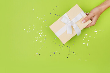 Women hands holding a gift or gift box decorated with confetti on a green table top view. Flat...
