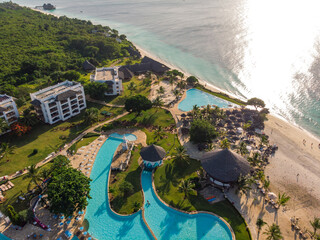 Aerial view of pool, umbrellas, sandy beach with green Palm trees. Coast of Indian ocean at sunset...
