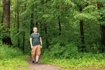 European young blond man in shorts and a gray T-shirt walks in the forest.