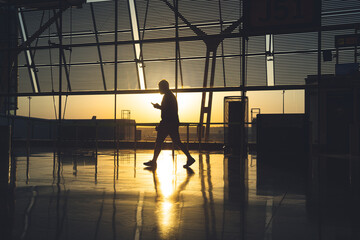 Naklejka na ściany i meble silhouette of an unrecognizable man staring at his mobile phone as he walks through the airport terminal at sunrise.