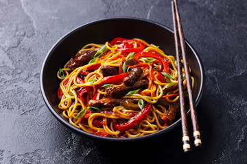 Stir fry noodles with vegetables and beef in black bowl. Slate background. Close up.