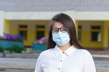 A schoolgirl in a medical mask in the background of the school. A high school student stands near the school. The girl in the antiviral mask.
