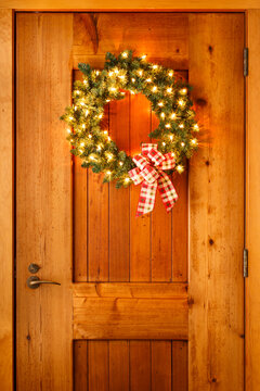 Beautiful lighted evergreen wreath with plaid fabric ribbon bow and lights hanging on wooden front door of house background. Simple, rustic, country style Christmas holiday home decorations.