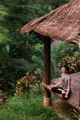 Young woman practicing yoga meditation sitting in easy pose in gazebo outdoors in green field on Bali. Unity with nature concept.