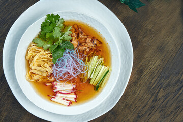 Cold korean guksu soup with rice noodles, cilantro and chicken in a white bowl on a wooden background.