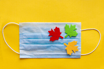 On the protective mask are colorful autumn maple leaves made of felt on a yellow background. Flat lay. Top view.