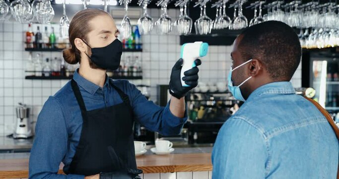 Caucasian male bartender in mask and gloves measuring temperature to African American client in cafe. Letting go customer in bar. Coronavirus concept. reopen at covid-19 quarantine. Mixed-races men.