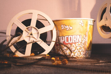 Old-school projector reel with uncoiled film is leaning against paper bucket of caramel popcorn in side warm light of lamp, front view. Traditional attributes of watching movies