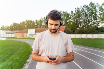 Man standing on the running track in day with smart phone and earphones - Adult caucasian male using mobile application to monitor training stats make a call send message - real people focus concept