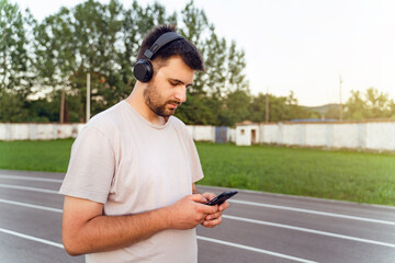 Fototapeta premium Man standing on the running track in day with smart phone and earphones - Adult caucasian male using mobile application to monitor training stats make a call send message - real people focus concept