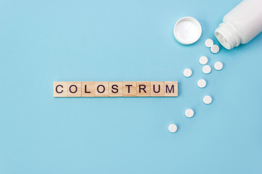 Word Colostrum. Wooden blocks with letters surrounded by pharmaceutical pills and plastic jar on blue background. Copy space, top view, layout