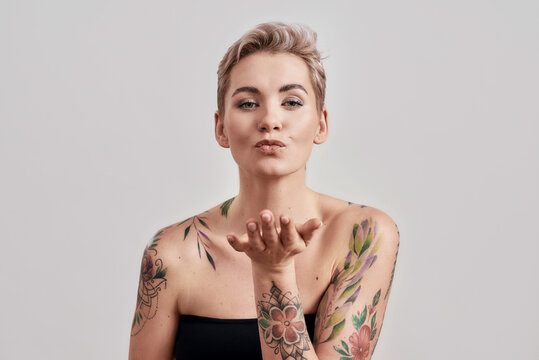 Portrait of a young attractive half naked tattooed woman with perfect skin blowing kiss to camera isolated over light background
