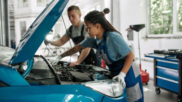 Genuine services. African american woman, professional female mechanic repairing car engine, tighten, screw with spanner while her colleague holding torch under car hood at service station