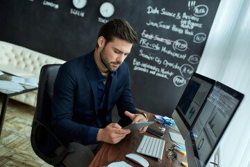 Business analyst at work. Young focused man sitting at his workplace in the office and using digital tablet. Analyzing statistical data, graphs and charts on pc screen