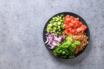 Salad of whole grain cereal spelt with vegetables, tomato, cucumber, greens on grey stone table with space for text.