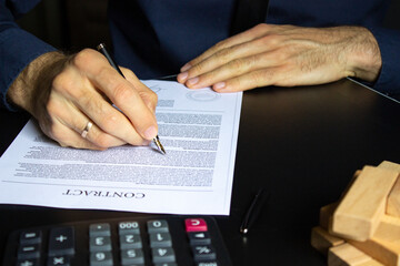 Hands of a businessman with a fountain pen over a contract, near a calculator and wooden blocks. The man checks the contract. Business concept.