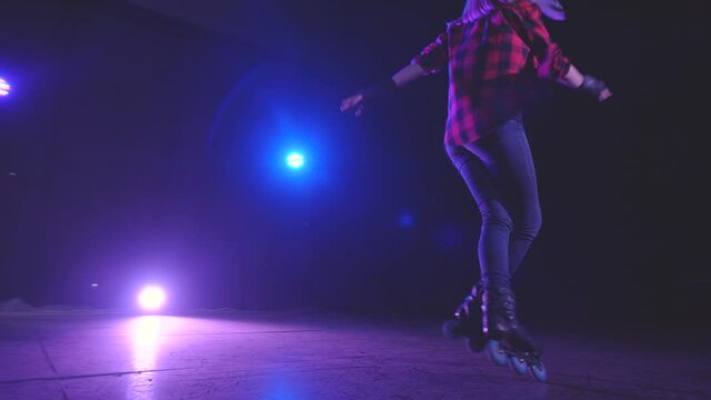 Close-up panoramic view of sporty pretty young girl spinning on roller skates in one place on front wheels under bright lights of spotlights on platform of abandoned building in accelerated shooting.