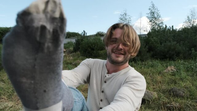 Funny blonde long-haired guy sits on park bench and puts sock on his leg. A guy in jeans and beige sweater dresses in the park.
