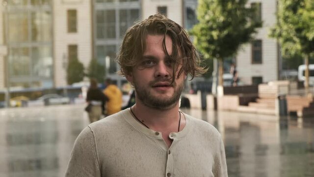 Serious guy with long wet blond hair got caught in rain and is looking at camera. Displeased young man portrait outdoors during day. The sun is shining into camera, the backlight is on the young man.