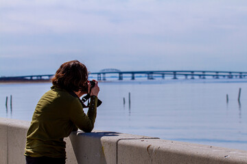 A young photographer is standing by a concrete wall and putting her elbows on the wall trying to take a picture of the Chesapeake Bay Bridge-Tunnel from distance. The bridge looks blurred.