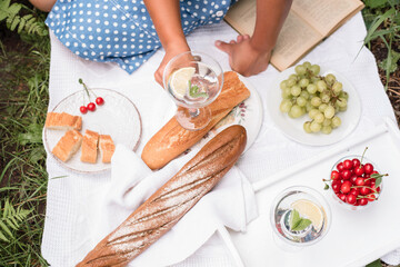 Summer picnic. A girl in a summer blue dress sits on a picnic in nature and drinks mojito. Picnic with baguettes, grapes, cherries and cocktail.