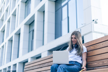 Beautiful woman with headphones working at laptop sit down on bench outside on a urban city street. Happy lady girl with wavy hair distance learning online education and shopping