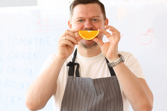 Vlogger Chef Holding Orange Slice Fruit Smile. Man Put Juicy Citrus Mouth On Face. Professional Cook Smiling On White Background. Home Culinary Concept. Cooking Male Looking At Camera Shot