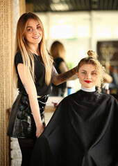 Blonde Hairstylist and Curly Hair Female Client. Beautiful Hairdresser Styling Wavy Strand and Bun for Young Woman. Two Girls in Beauty Salon. Styling Hair Curls for Customer Looking at Camera Shot