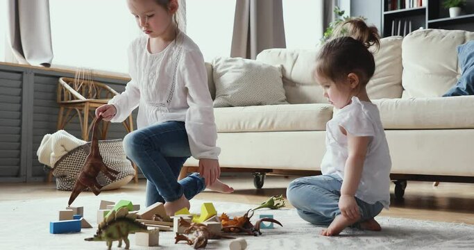 Two little preschool daughters children sitting on floor, playing toys in living room. Small cute girl ruining wooden building construction, having fun with elderly sister at home, making her upset.
