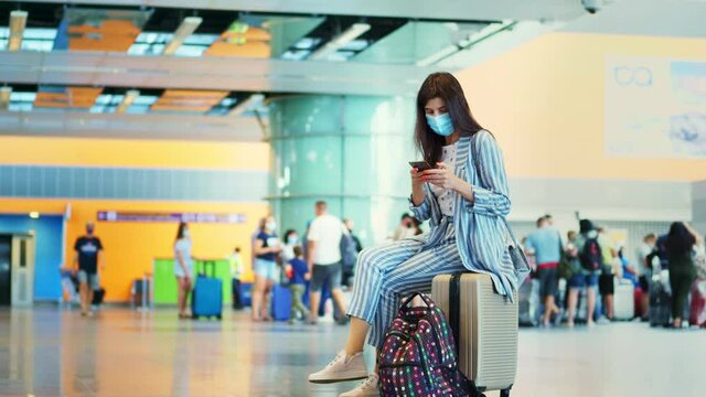 woman in mask, sitting on a suitcase, luggage, against background of air passengers crowd, at airport. checking flight timetable using mobile. Trip after coronavirus epidemic.