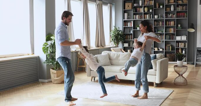 Full length active young couple holding little daughters in hands, twisting barefoot on warm floor indoors. Happy parents having fun, dancing to energetic music with funny small kids sisters at home.