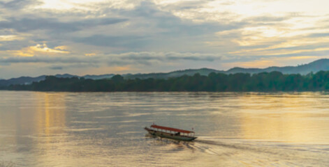 Blur background of a boat with a boatman in the middle of the Mekong River in the evening. The sky is cloudy and the orange light of the setting sun with a backdrop of mountains and lush vegetation.