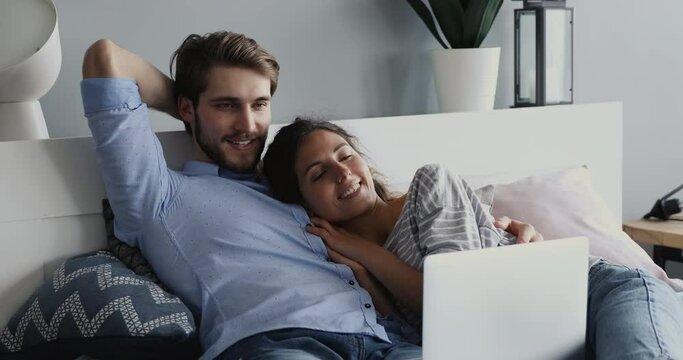 Happy loving married couple lying on cozy bed watching funny movies or comedian series on laptop indoors. Smiling relaxed affectionate spouses enjoying spending calm weekend holiday time together.