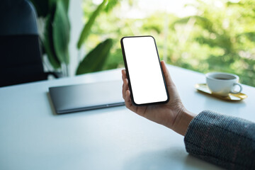 Mockup image of a business woman holding mobile phone with blank white desktop screen with laptop on the table