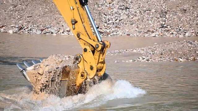 Excavator bucket close up. Excavator digging river rocks sand and pebbles. Heavy industry and environment. Ecological disaster. Machine dredging river canal. Dam building. Slow motion, static shot