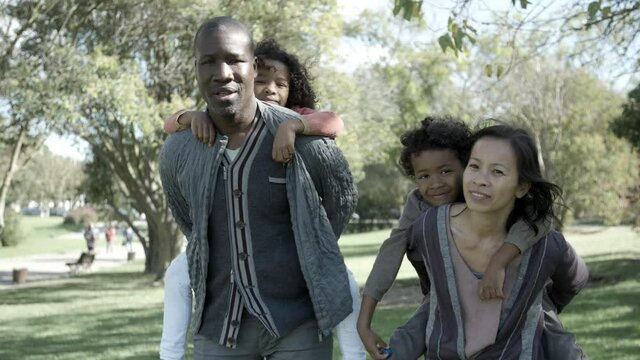 Young Asian mother, African American father holding kids on back and smiling. Mom and dad standing in city park with son and daughter hugging them. Front view. Family, nature and weekend concept