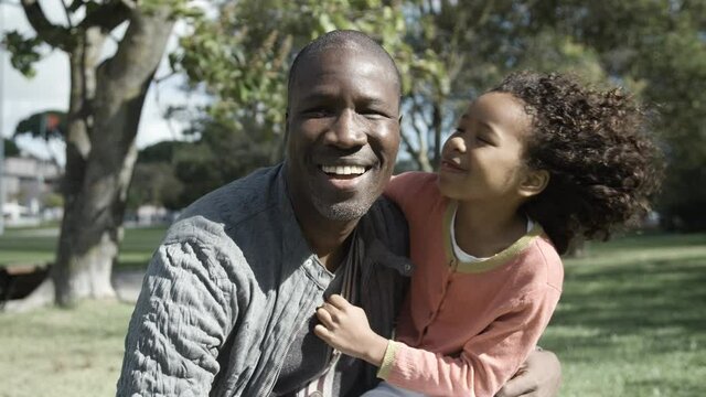 Cute girl kissing and hugging her African American father in park. Bald dad smiling and looking at camera. Front view. Medium shot. Static camera. Family, holiday and weekend concept
