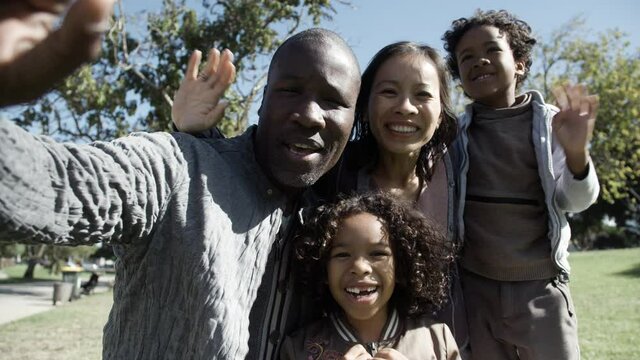 Mixed-race family waving, greeting, smiling and looking at camera. African American father holding device, Asian mom and kids speaking with someone. Family, video chat and holiday concept