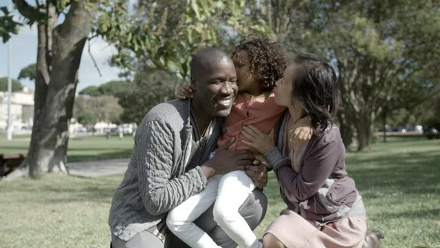 Cute girl sitting on dad knee, kissing and hugging parents. Asian mom and African American father embracing daughter and smiling. Front view. Medium shot. Static camera. Family and weekend concept