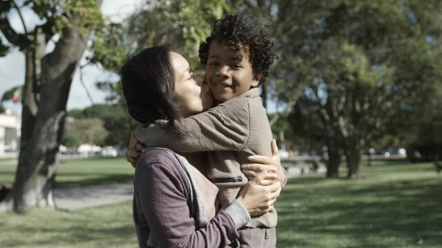 Cute mixed-race boy kissing and hugging Asian mother in park. Mom standing, holding kid, smiling, talking and closing eyes. Front view. Medium shot. Static camera. Family, holiday and weekend concept