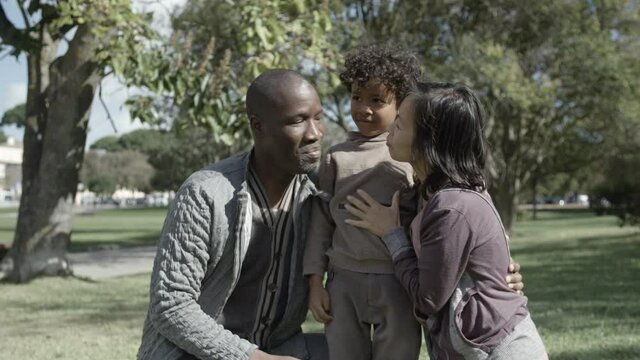Cute boy kissing and hugging Asian mom and African American dad in park. Parents embracing kid, smiling and posing to camera. Front view. Medium shot. Static camera. Family and holiday concept